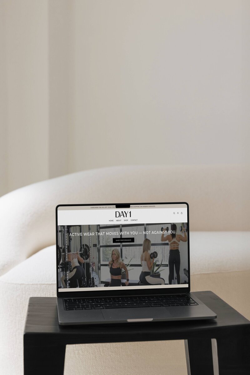 Day 1 Fitness website showcased on an open Macbook Pro laptop sitting on top a black table in front of a cream couch