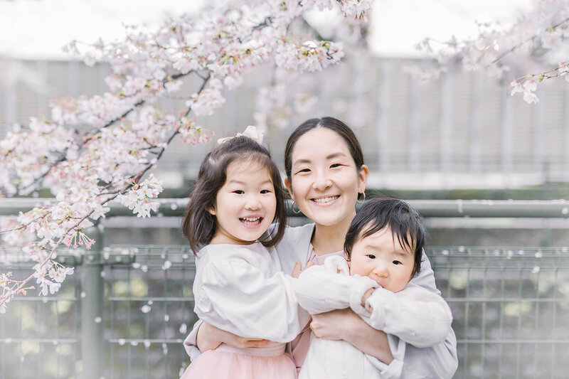 桜の花をバックに、女の子と男の子の赤ちゃんを抱っこして笑顔を見せている女性の家族写真。明るくて優しい雰囲気。