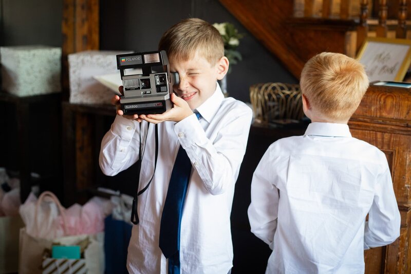 Inside a restored Pennsylvania farmhouse, two young boys in dress shirts and ties share a playful moment. One squints through a vintage Polaroid camera while the other faces away, surrounded by wooden beams, wrapped gifts, and soft natural light. It’s a slice of real-life charm during a Philadelphia area wedding prep.