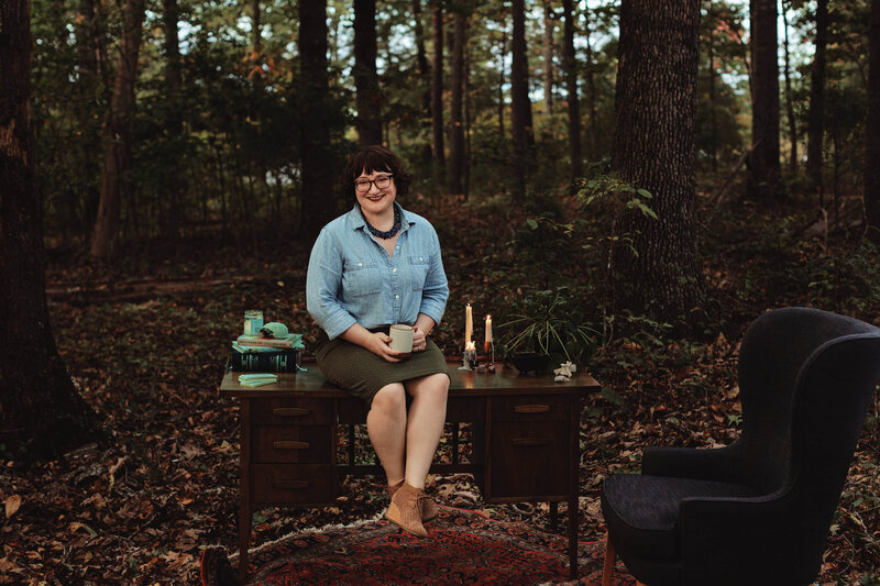 Energy worker Ash Stinson sits on a wooden desk holding a coffee mug set in the forest