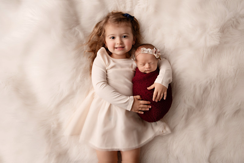 Little girl holding her baby sister on white rug taken by Detroit Newborn Photographer Kat Figlak