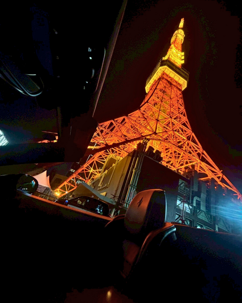 Brightly lit Tokyo Tower viewed from the interior of a Ferrari convertible at night, with the tower’s glow casting reflections on the car’s surfaces.