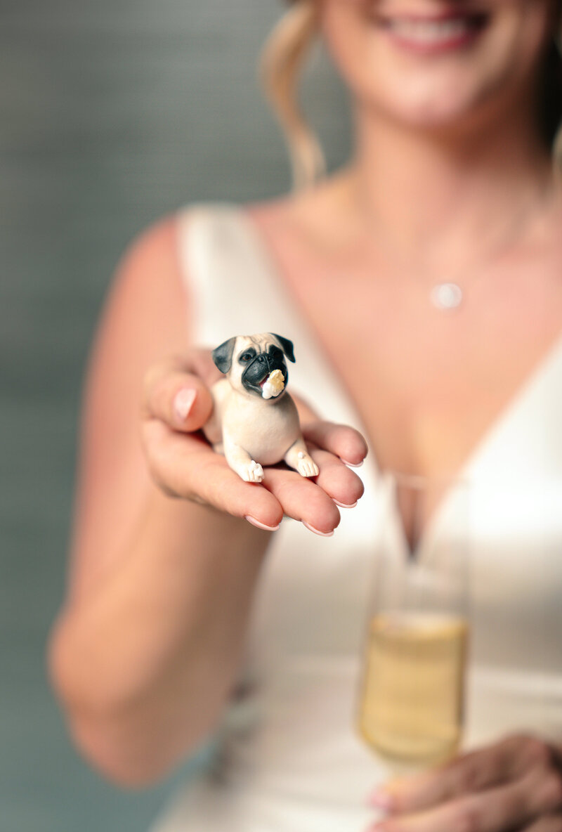 A person in a white dress is holding a small figurine of a pug in their hand. They are also holding a glass of champagne in the other hand. The background is blurred.