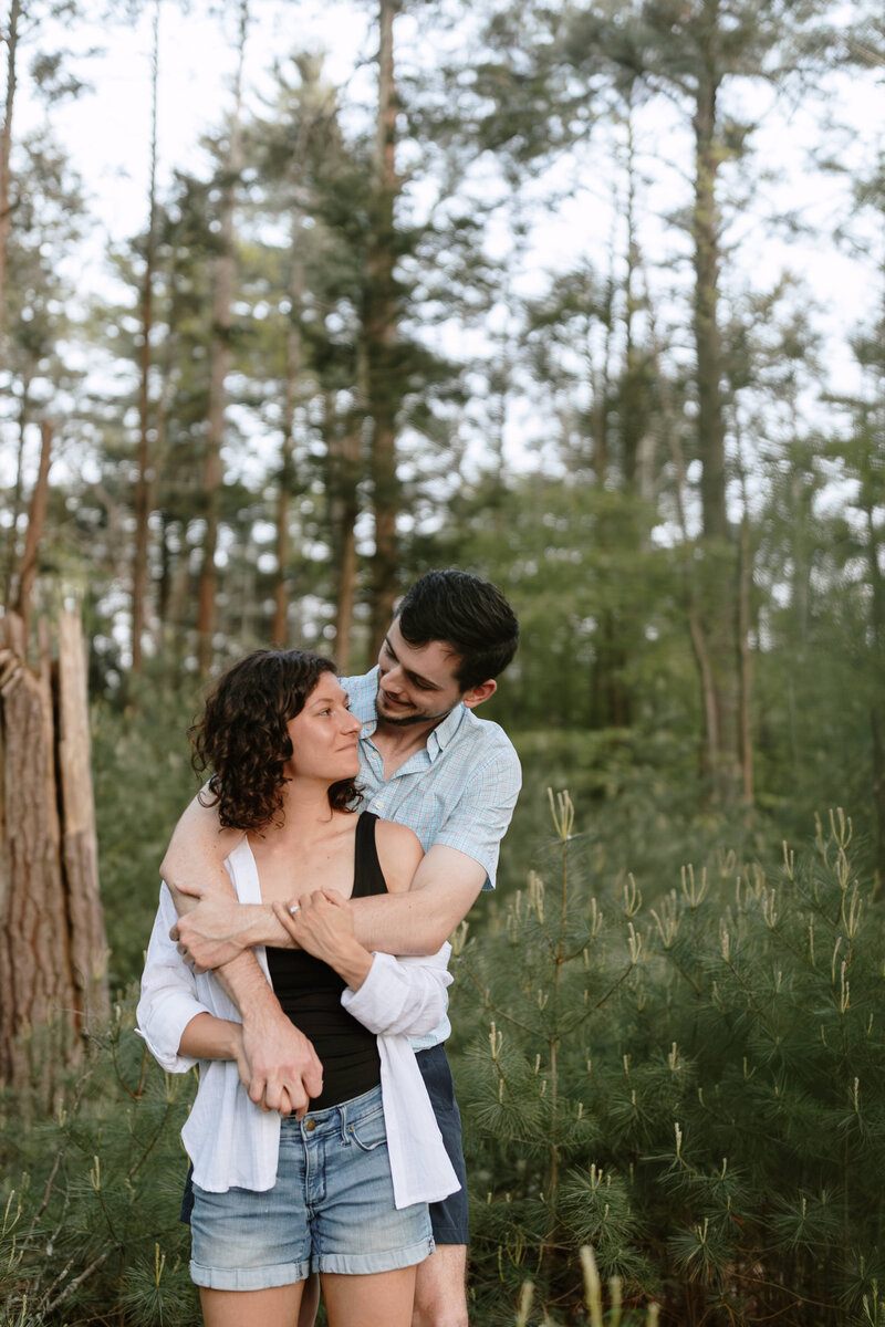 A couple embraces in a wooded area, standing among tall trees and greenery,