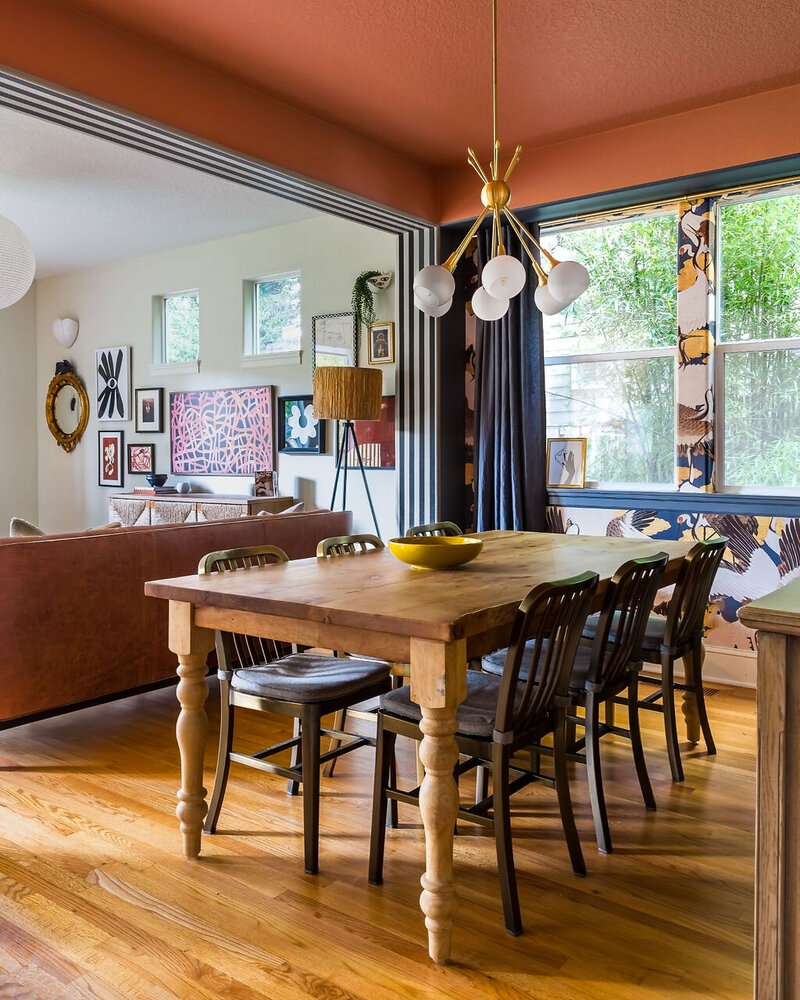A wooden dining room table with a decorative lamp handing above it and a peek into the living room behind it with colorful art on the wall, representing a maximalist interior design.
