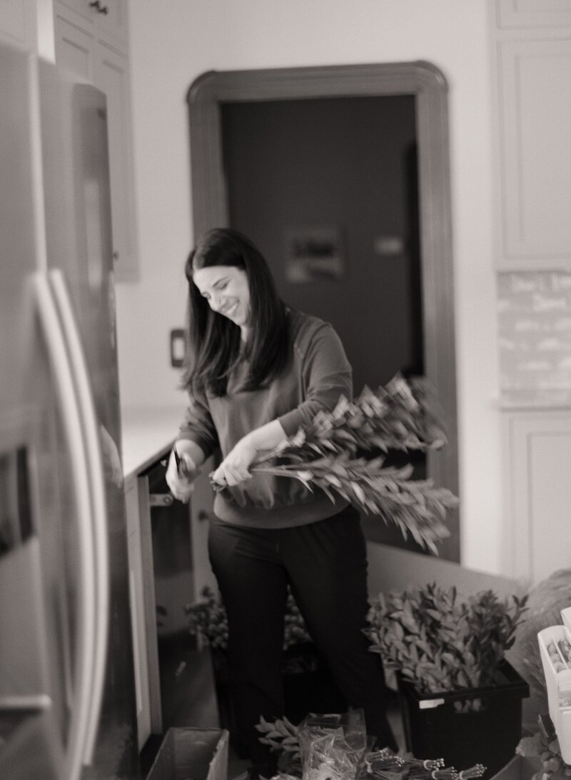 Soft-focus black-and-white image of a smiling florist holding stems of greenery in one hand and snipping them with clippers at a kitchen counter.