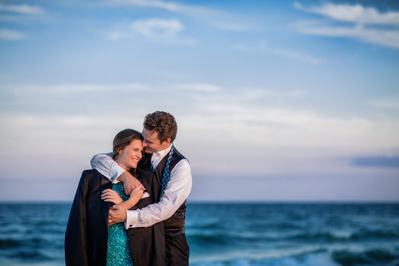 A couple walking hand in hand on a sandy beach at the New Jersey shore during their summer engagement session, snuggling and enjoying the ocean breeze.