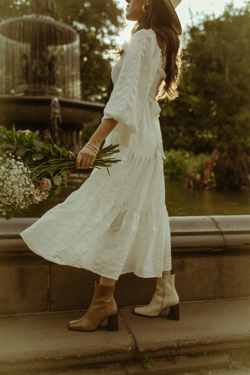 A woman  carrying a bunch of blush pink roses in motion with the wind making her white dress and the flowers move