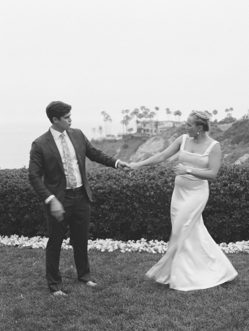 A bride and groom in la jolla having a fun private dance. They are standing on some grass infront of a hedge with a view of the ocean and some palm trees behind them. They are standing a few feet apart looking at eachother and looks like they are spinning toward eachother