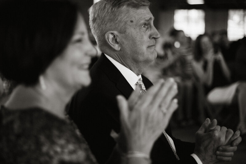 Candid black and white photo of parents clapping joyfully during a wedding ceremony, captured by Rainstorm Photo & Video in Michigan City, Indiana.