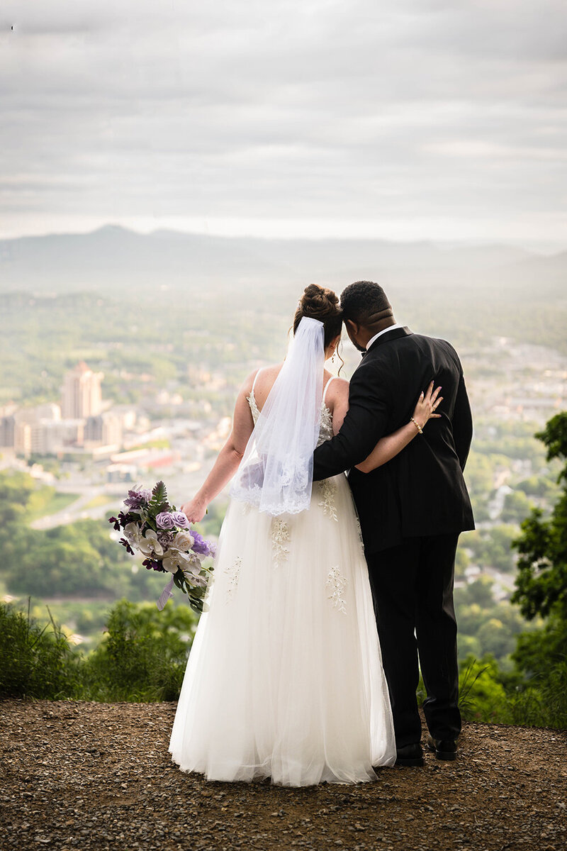 A couple on their Virginia elopement day wrap their arms around one another and enjoy the view of the Downtown Roanoke skyline and the Blue Ridge Mountains.