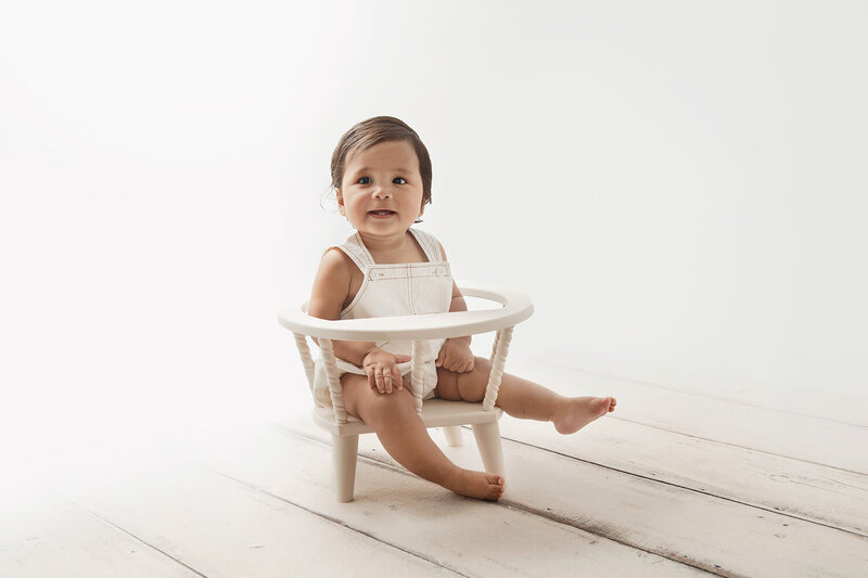 A young toddler sits in a baby chair in white overalls in a studio