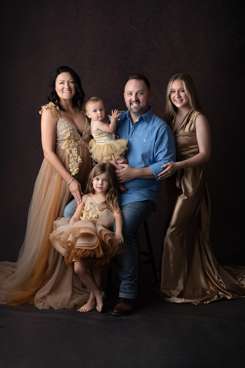 Family of five poses together in coordinated gold and neutral tones, with the mother and daughters wearing flowing gowns and the father in a blue shirt and jeans. Captured in a timeless fine art style by Dallas Family Photographer Lindsay Walden.