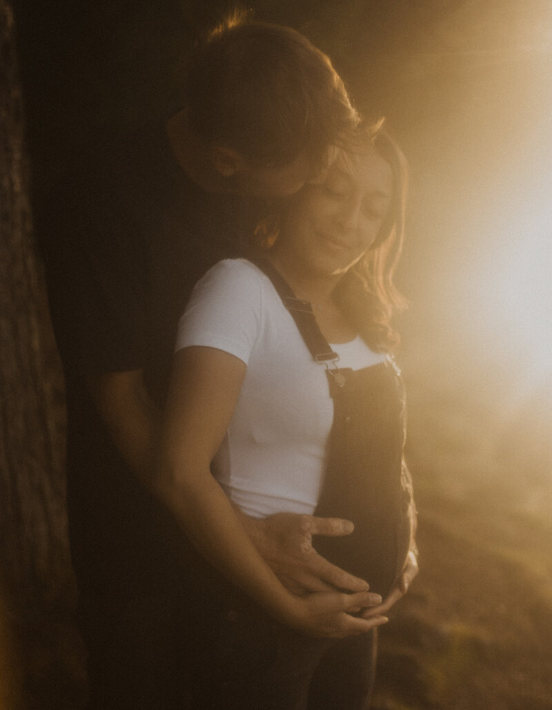 A man stands behind his pregnant wife and holds her belly. He gives her a kiss and they both have their eyes closed and smile. It is golden light.