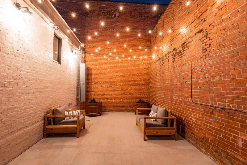 A narrow outdoor patio with string lights overhead, two wooden sofas with cushions on either side, potted plants at the far end, and surrounded by tall brick walls.