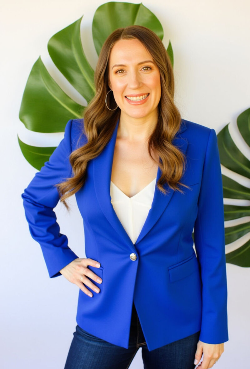 Stacey from Xanthe Bookkeeping smiling in a cobalt blue blazer, standing with her hand on her hip in front of sculptural Monstera leaves.