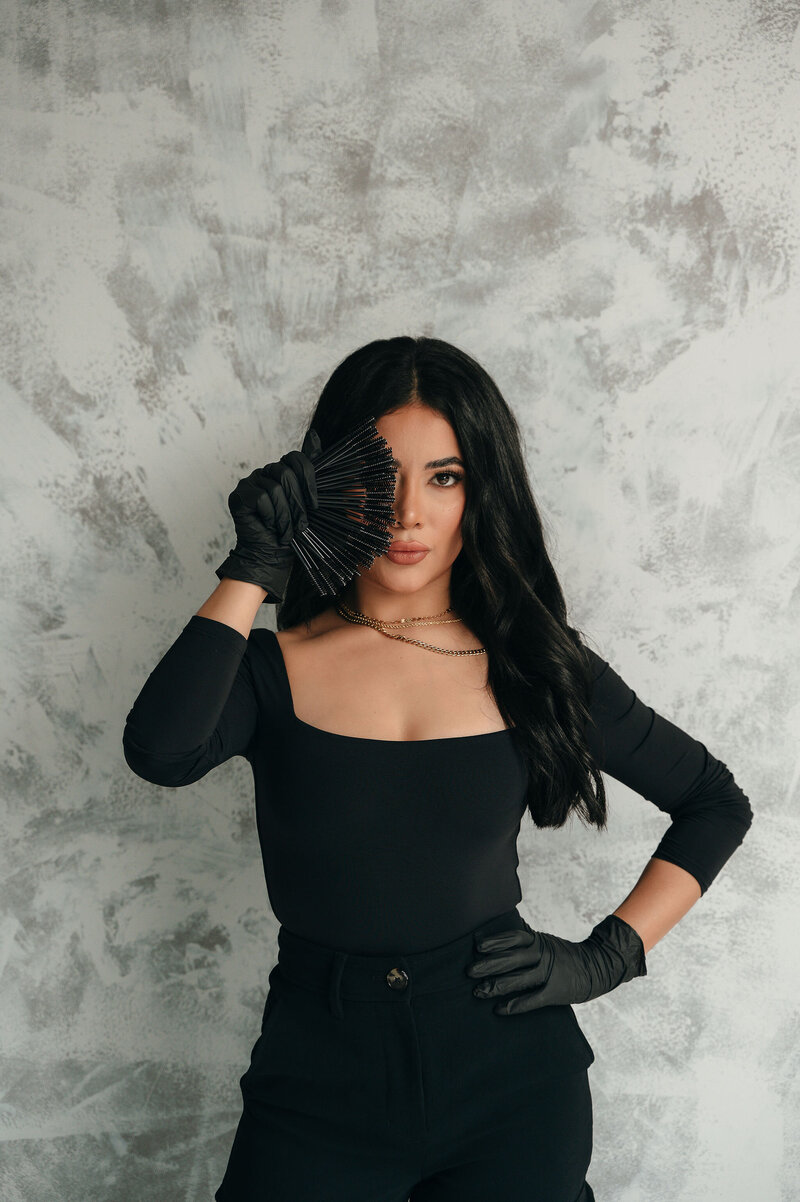 A woman in a white shirt and jeans paints a pink flower on a white wall, holding a palette. A mirror reflects her image, suggesting focus and creativity.