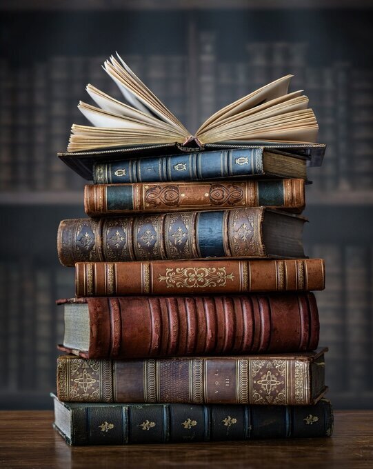 Eight antique books with colorful leather spines are stacked on a wooden table in a library. The ninth book lies on top with its pages open.