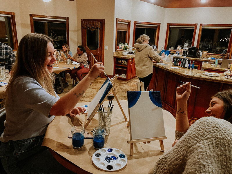 Women sit at a table in a kitchen painting on small easels. 
