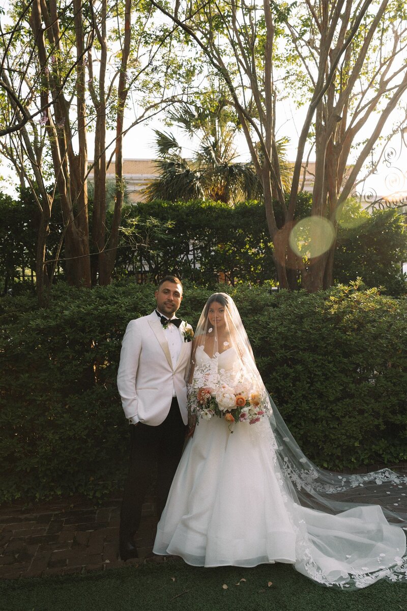 bride and groom stand together in front of green hedges at William Aiken house in charleston. while looking at the camera with the bride under her veil.