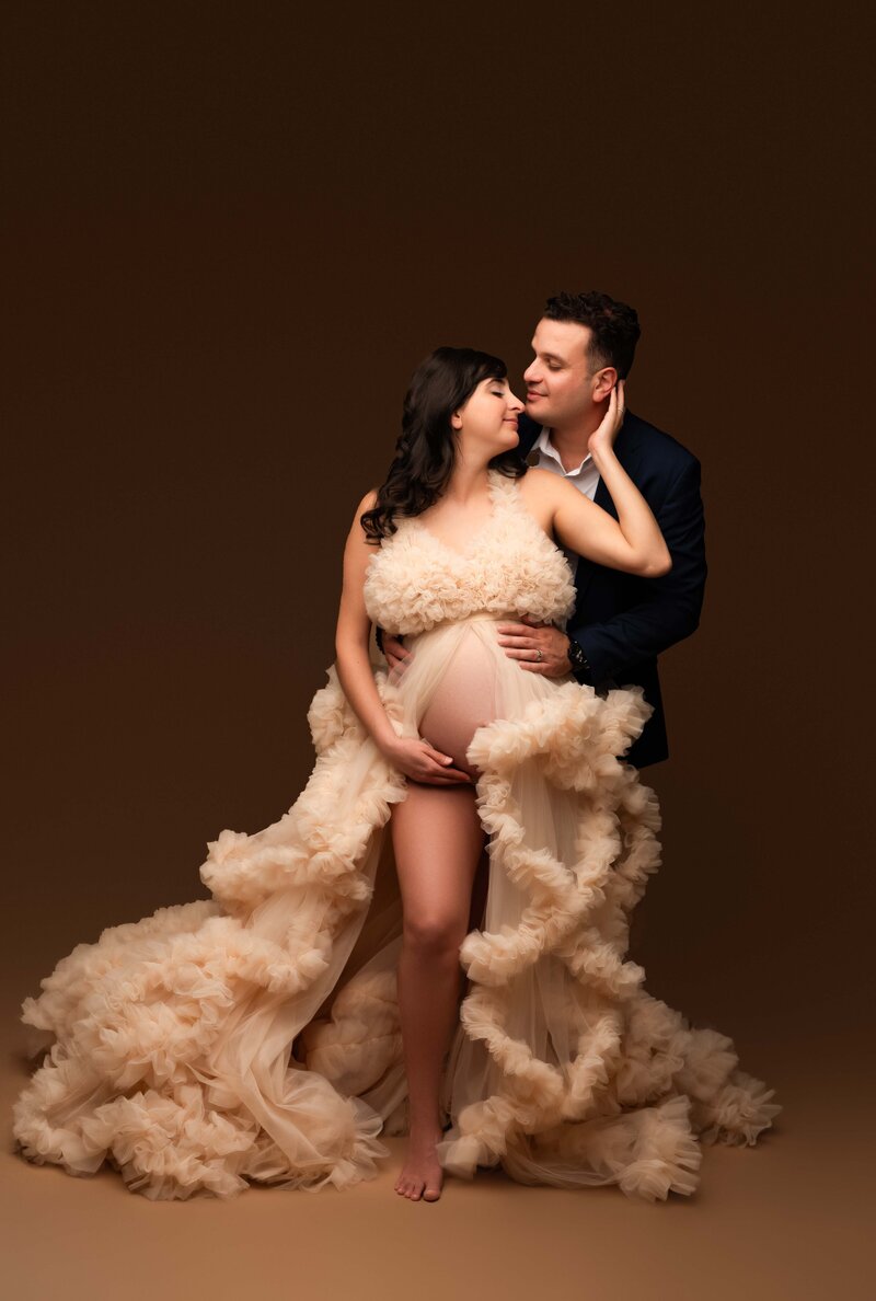 Pregnant mother and father to be in nude tulle gown and navy suit looking at each other touching noses