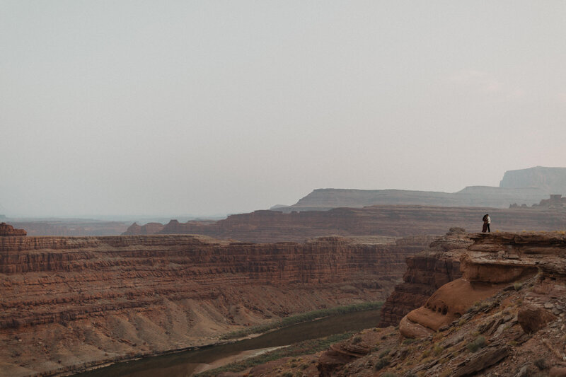 A couple stands next to a cliff overlooking the Colorado River during their offroading engagement session with a Jeep near Moab Utah