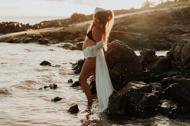 Pregnant woman in a black bikini and soft white cover up draping off her shoulder  poses on a beach at sunset. Her side profile is facing the camera