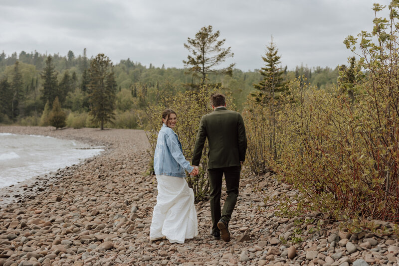 bride and groom walking along a beach on the north shore, bride is wearing a denim jacket and is looking back and smiling.