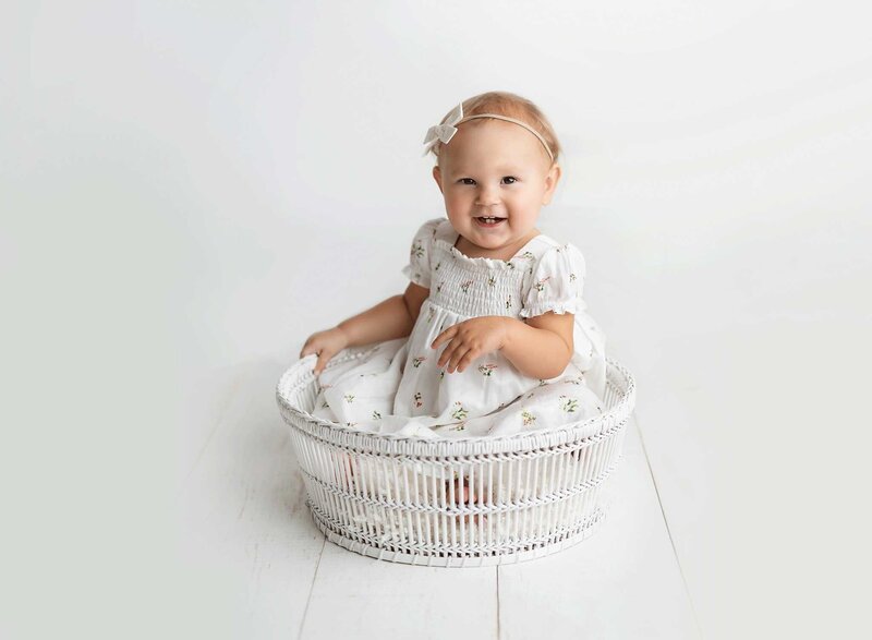 A young toddler girl in a white dress and bow sits in a white wicker bucket