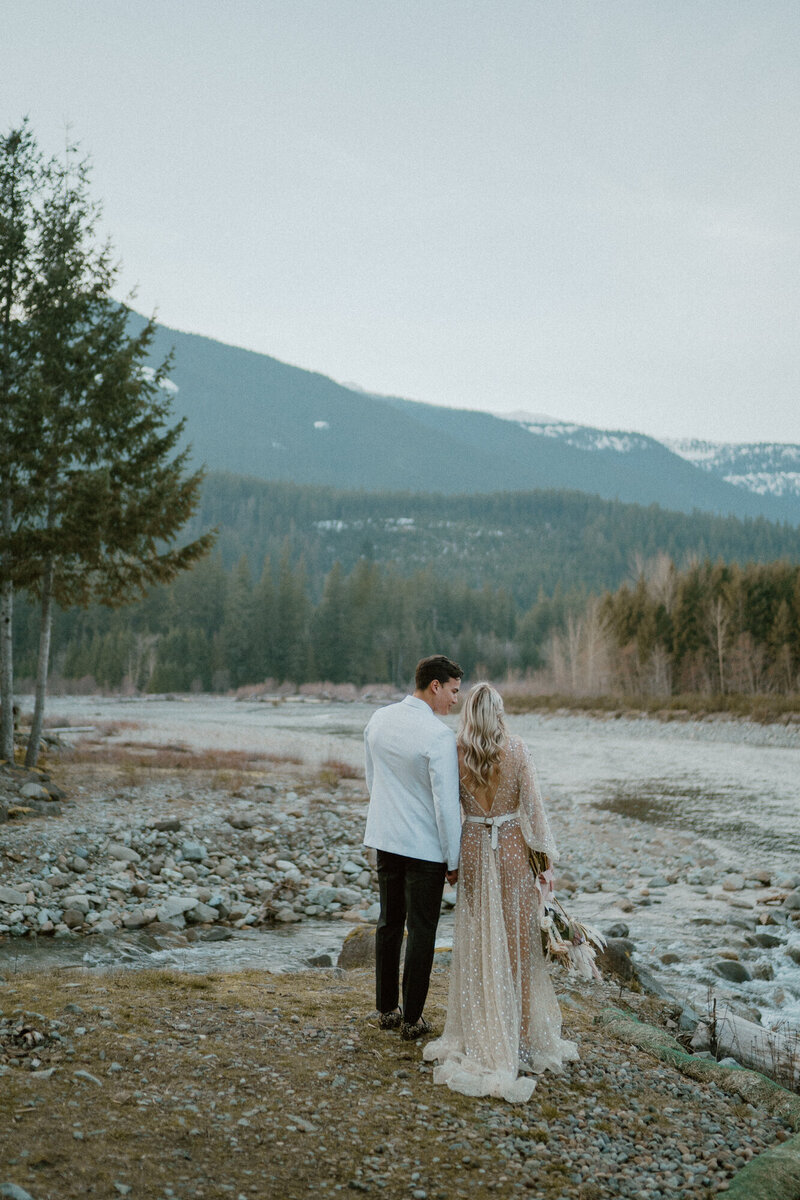 bride and groom holding hands near river with backs to camera