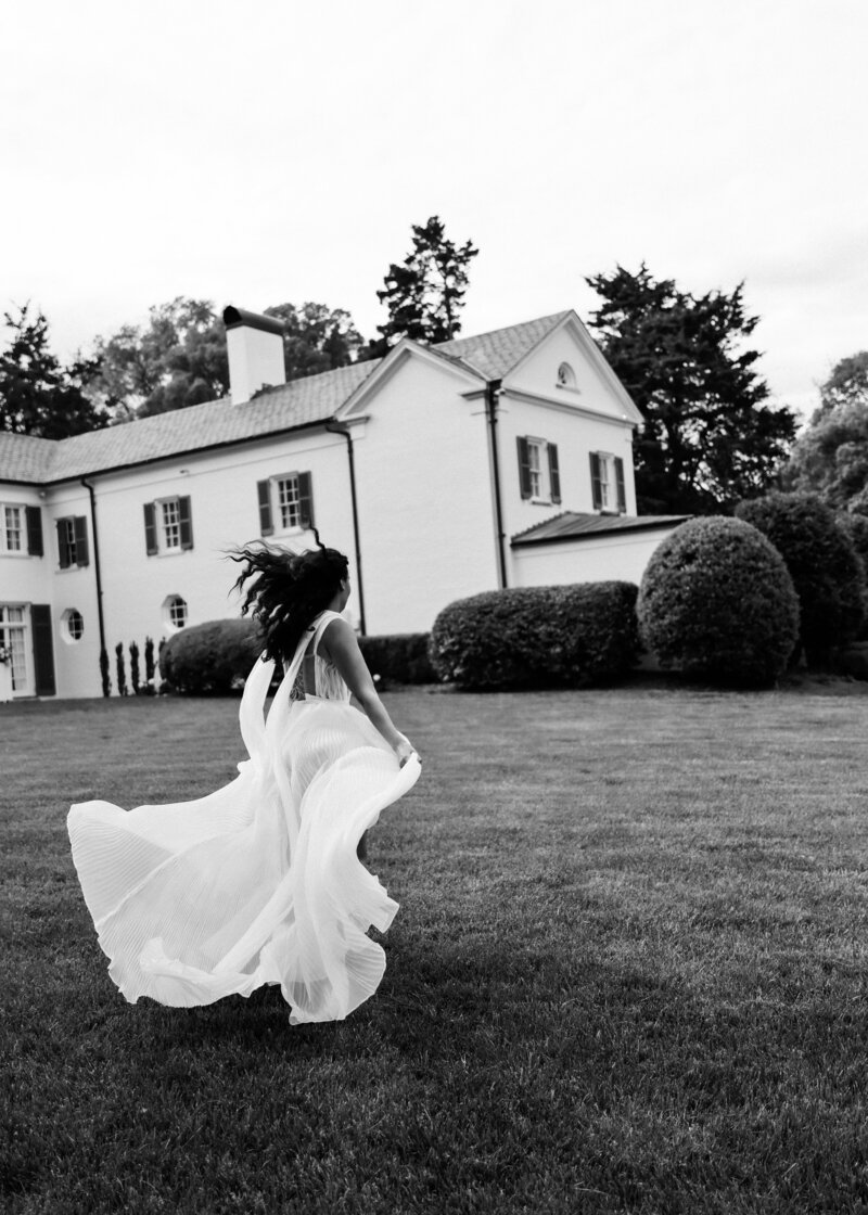 Julia stands by the window at Boxwood Estate, gazing out as she prepares for her wedding day. She wears a beautiful wedding dress with intricate details and a flowing veil. The soft light filters through the window, creating a serene and intimate atmosphere. Her calm expression reflects the anticipation of the special day ahead, with the elegant interior of the estate adding to the moment’s beauty.
