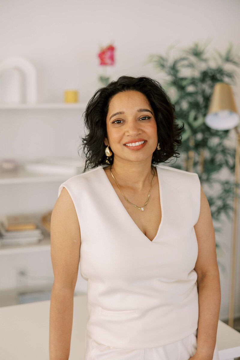 Woman leaning casually on a table, wearing an all white outfit and smiling at the camera