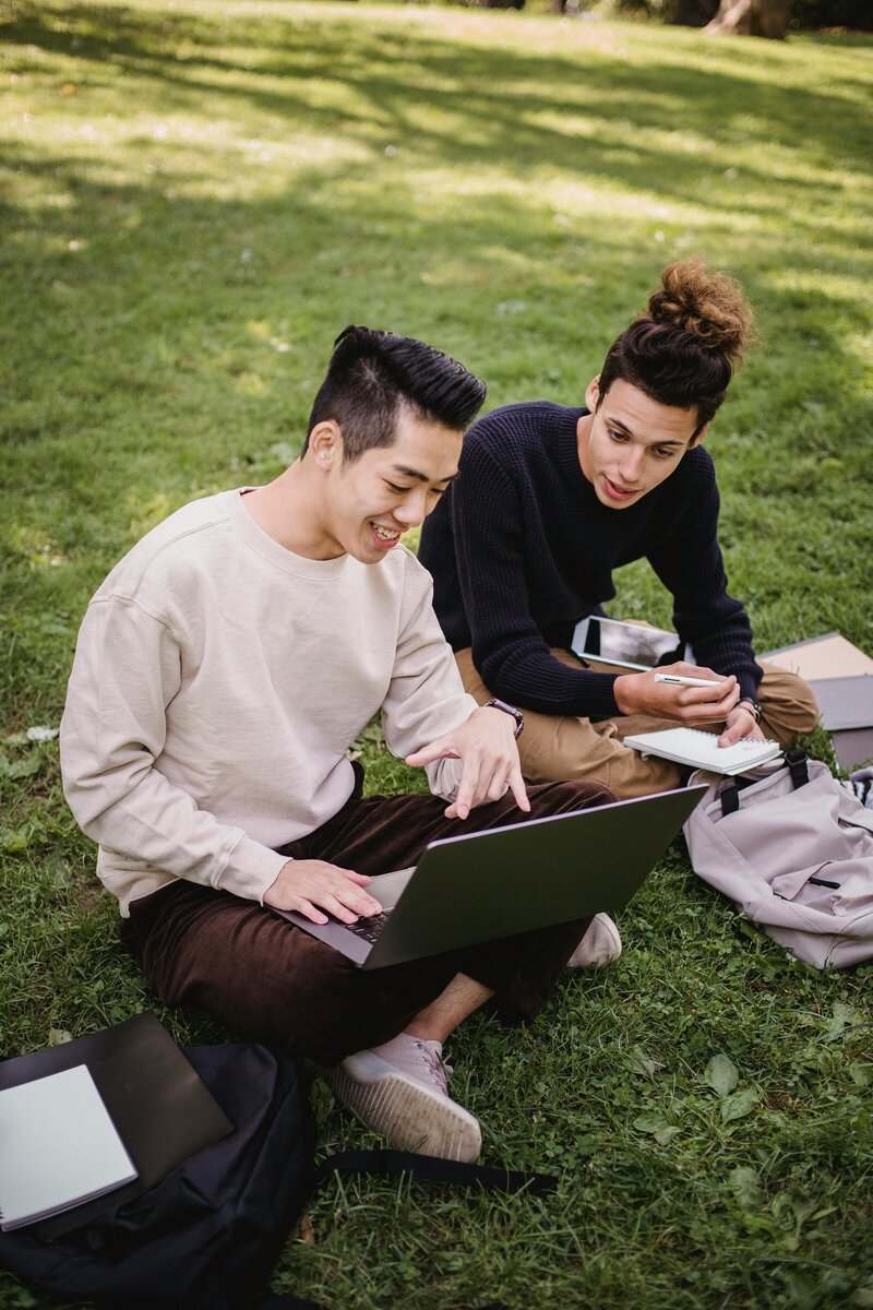 Two friends sitting on the grass, working together on a laptop and taking notes in a relaxed outdoor setting