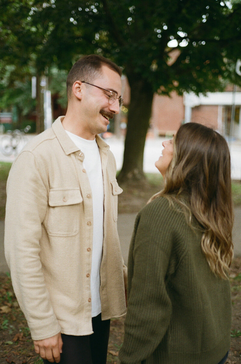 A man and woman stand outdoors, facing each other and smiling. The man wears a beige jacket, and the woman dons a green sweater, capturing a perfect moment. With trees and a building in the background, it's an ideal scene for an Ontario destination wedding photographer.