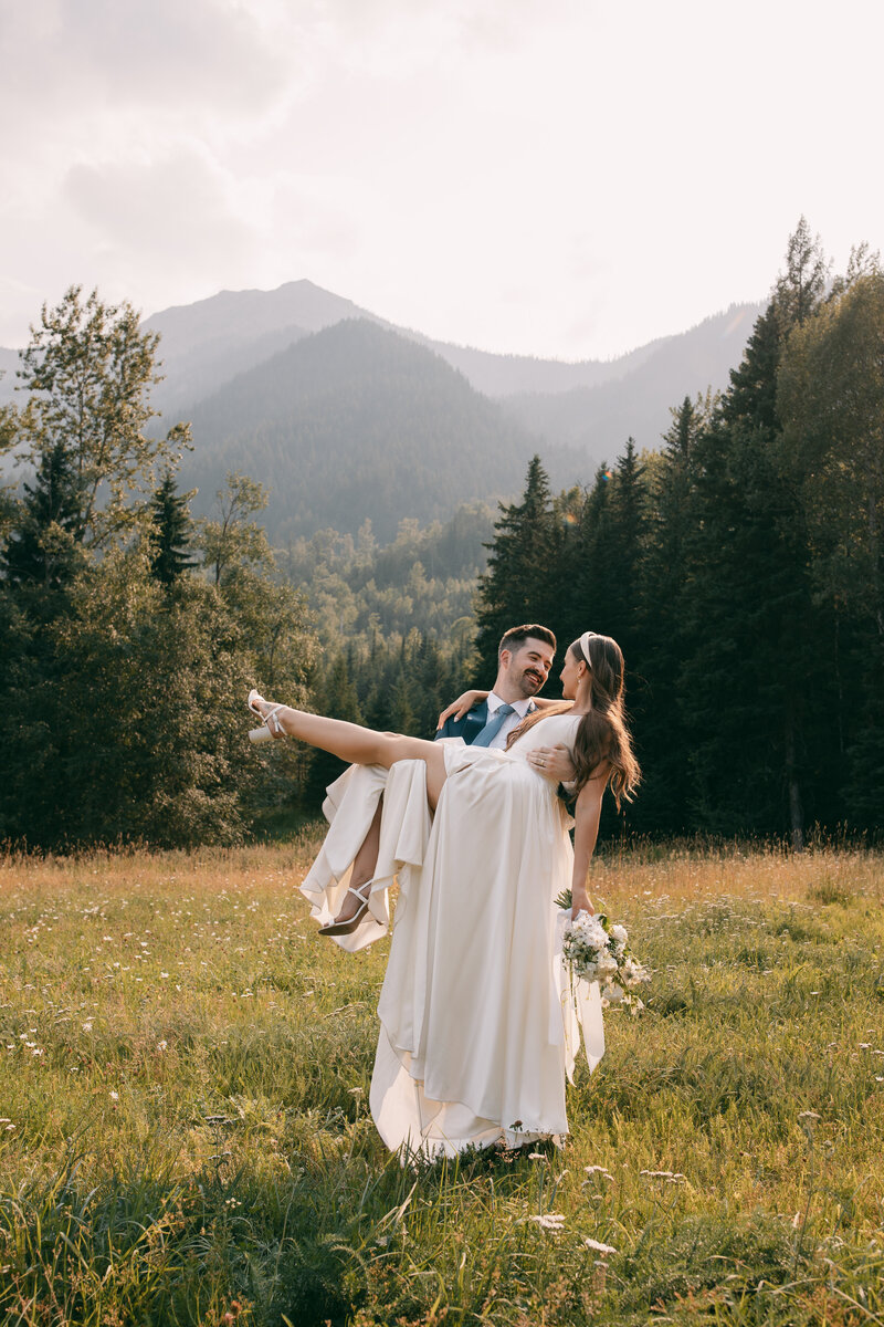 A newlywed couple with the groom holding the bride in his arms with a mountain backdrop