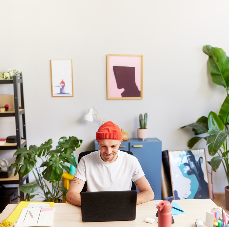 Creative professional in a red beanie focused on his laptop in a vibrant, modern home office decorated with plants, framed art, and a variety of desk accessories.