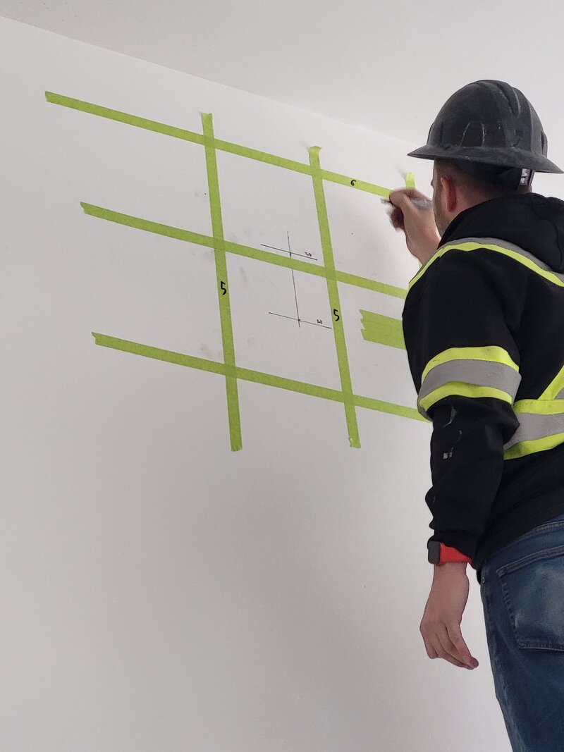 An Operator is writing the depth at which each object in the concrete was found on a wall within a grid made of green tape.