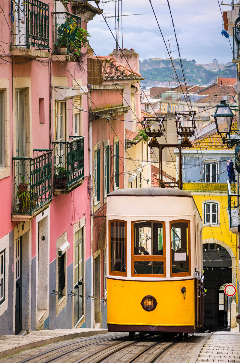 A classic yellow tram travels up a steep, narrow street lined with colorful buildings in Lisbon. Pink and yellow facades with balconies and hanging laundry frame the scene, while overhead cables crisscross above the tram as it ascends the hill.
