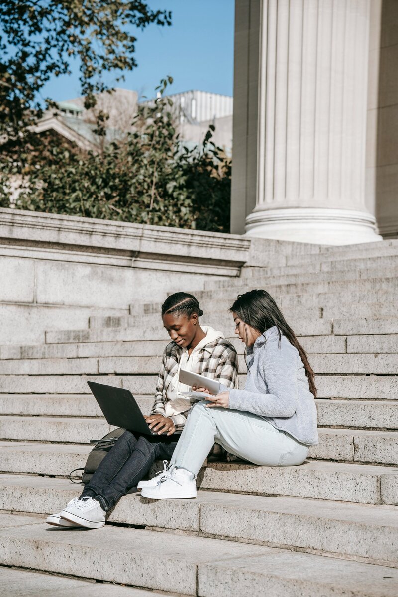 Two friends sitting on outdoor stone steps using a laptop and notebook while studying in the sun
