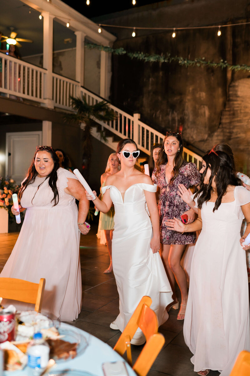 A bride in a white gown and heart-shaped sunglasses walks with bridesmaids in light pink dresses, holding glow sticks, at an outdoor wedding reception with string lights and a staircase in the background.