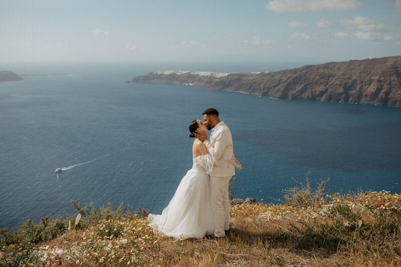 Bride and groom embracing on a cliffside in Santorini with wildflowers in the foreground and the Aegean Sea in the background.