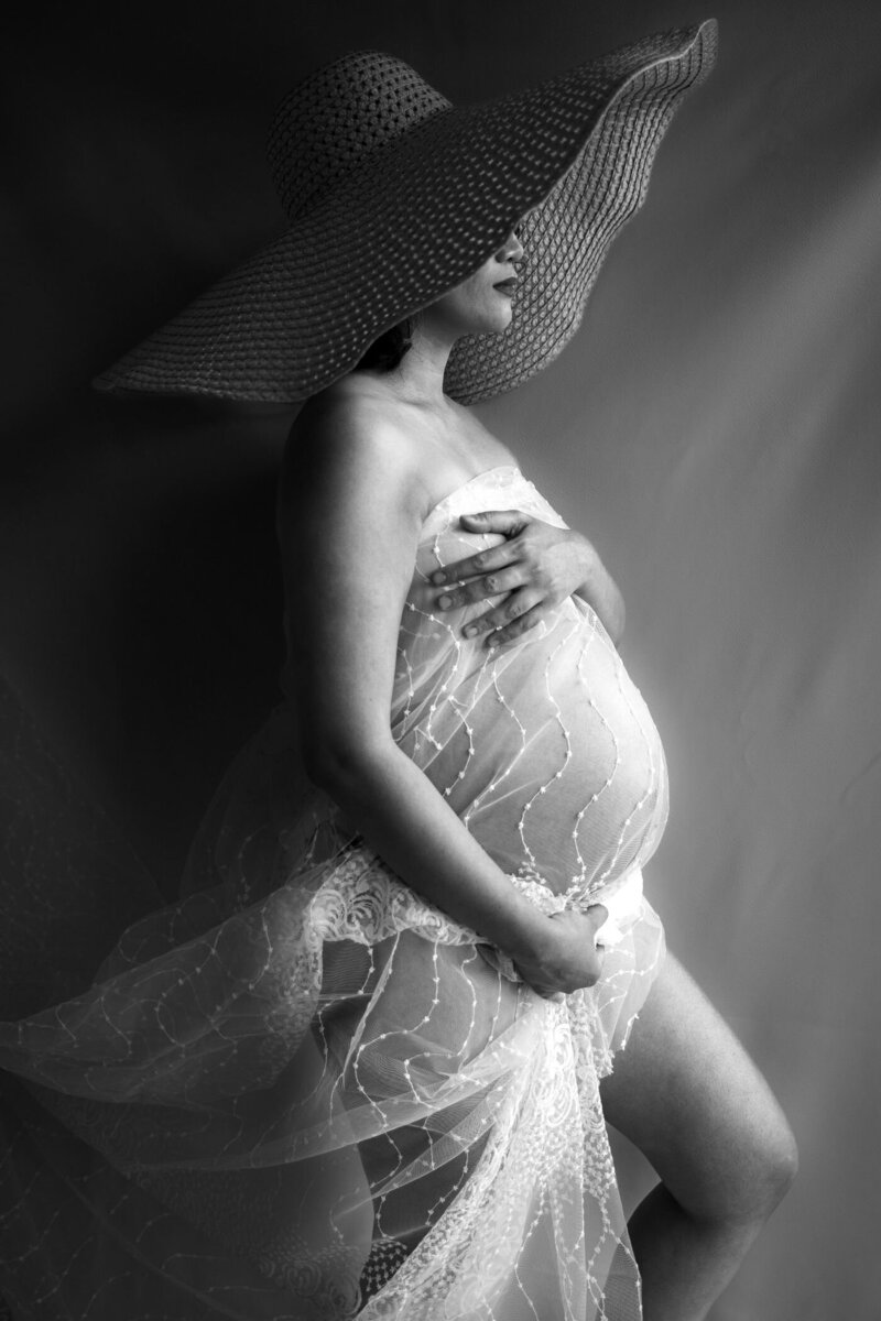 Black-and-white artistic side profile of a pregnant woman wearing a dramatic wide-brimmed hat and sheer lace fabric, highlighting the elegant curve of her baby bump.