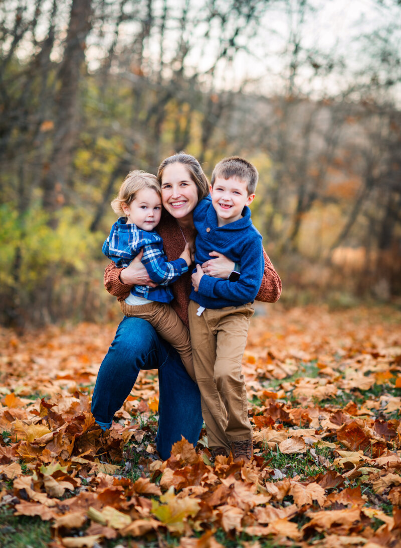 A mother holding her two children, one in each arm, against a backdrop of autumn leaves by Leslie Q Photography