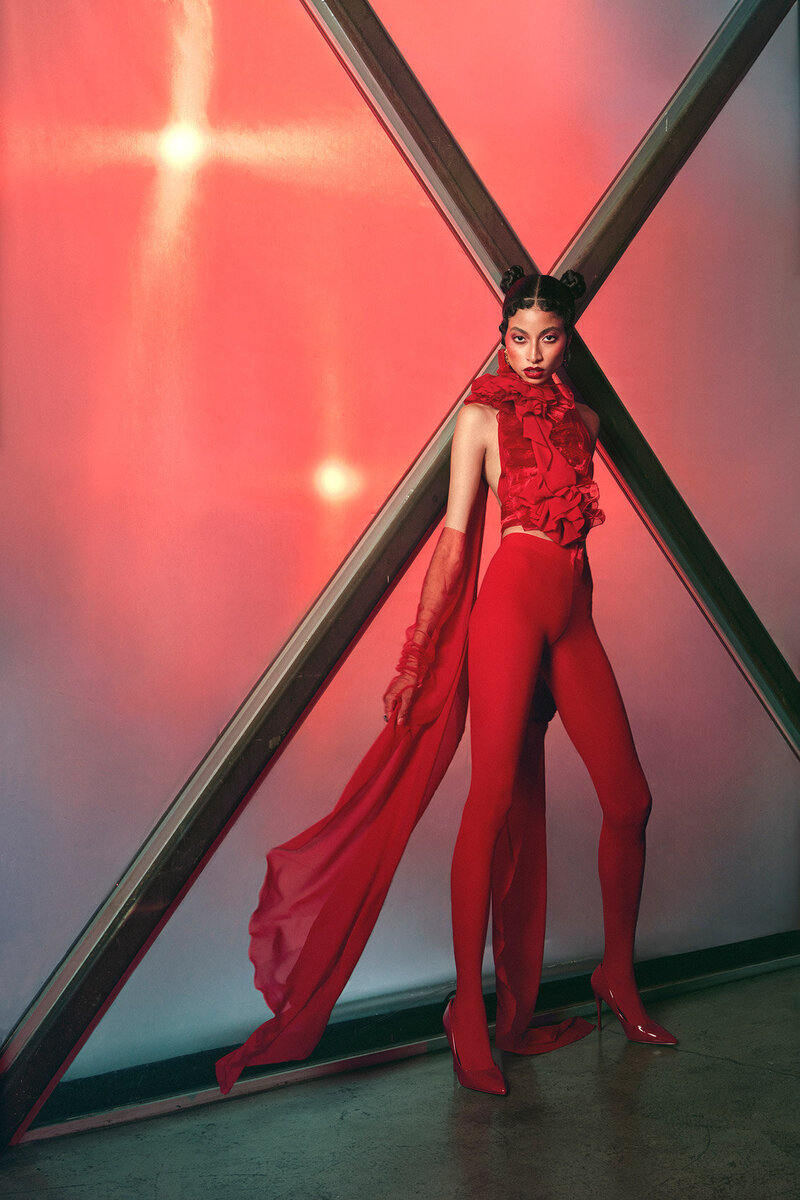 Striking portrait of Nic Shiya in a bold red harness outfit, set against a red backdrop, captured by Ylva Erevall.
