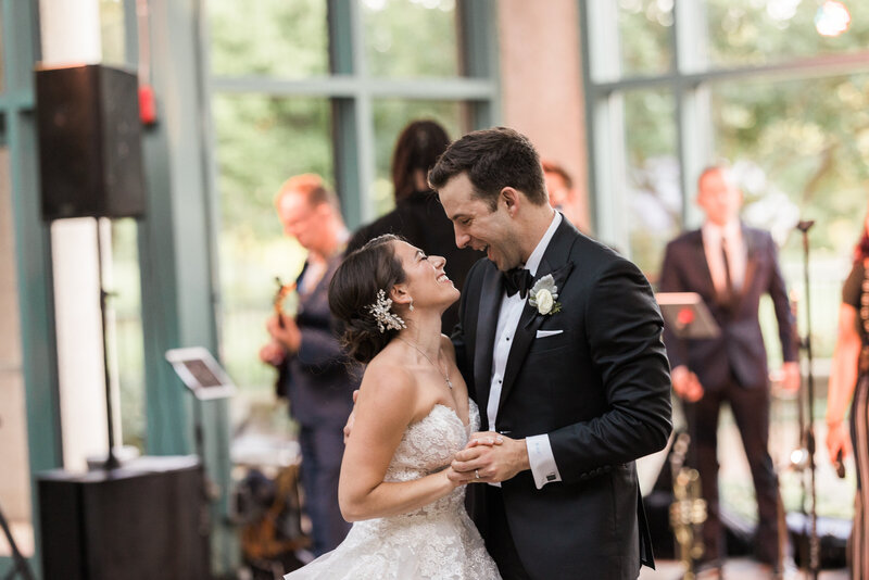 white groom in black tux on a dance floor with white brunette bride in strapless white gown laughing at one another during their first dance with the wedding band in the background