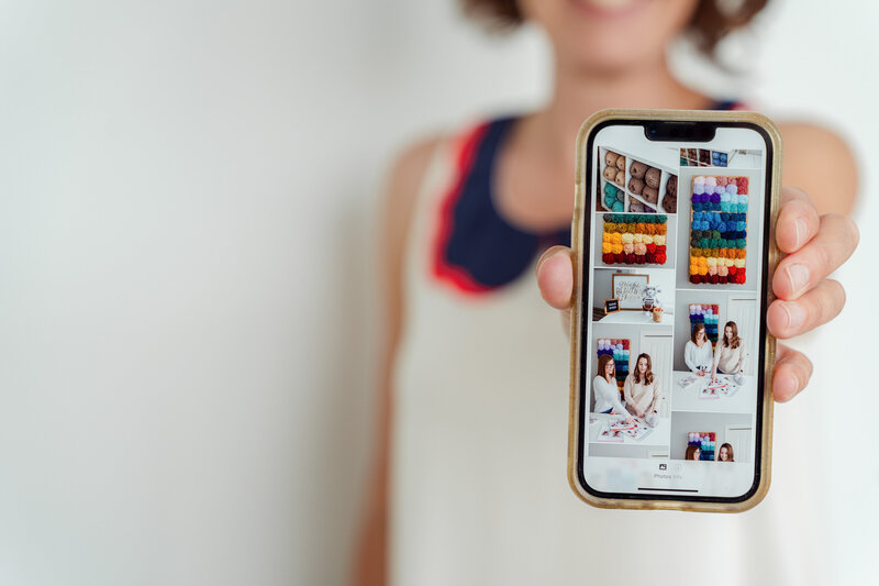Kate Daniel Photography holding a mobile phone in front of her that shows Calgary brand photography images of Double Dutch Crochet Co. Kate is blurred out in the background.