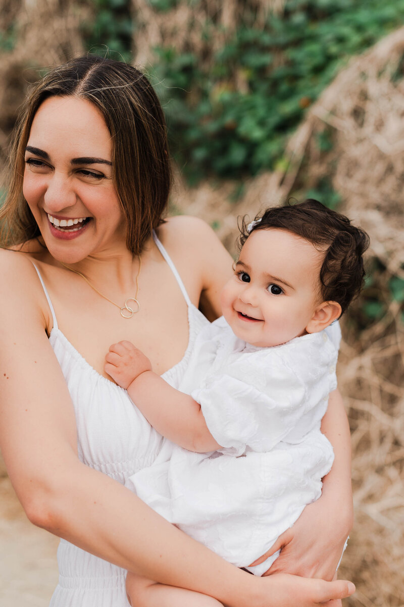 Mom laughing while holding her baby and being photographed by a Los Angeles family photographer.