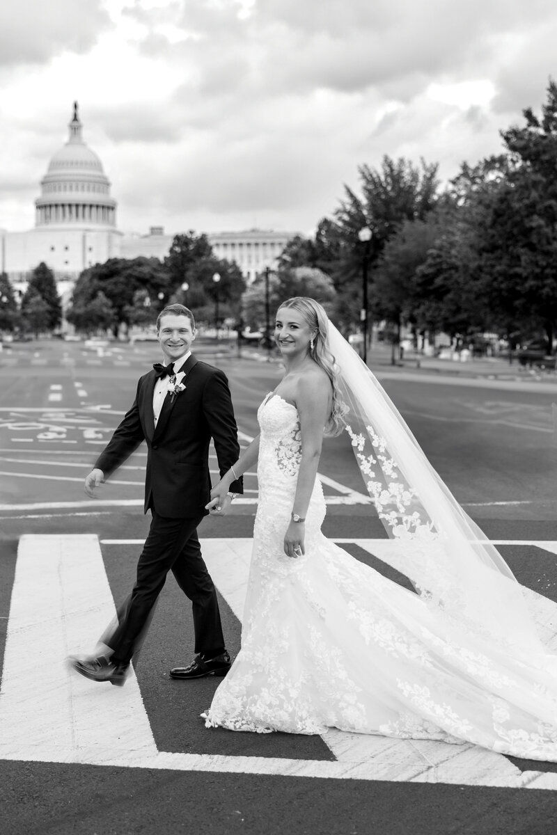 A bride and groom, smiling and holding hands, walk across a crosswalk with the U.S. Capitol building in the background. The bride wears a long, lacy veil and gown; the groom is in a tuxedo. The scene is in black and white.