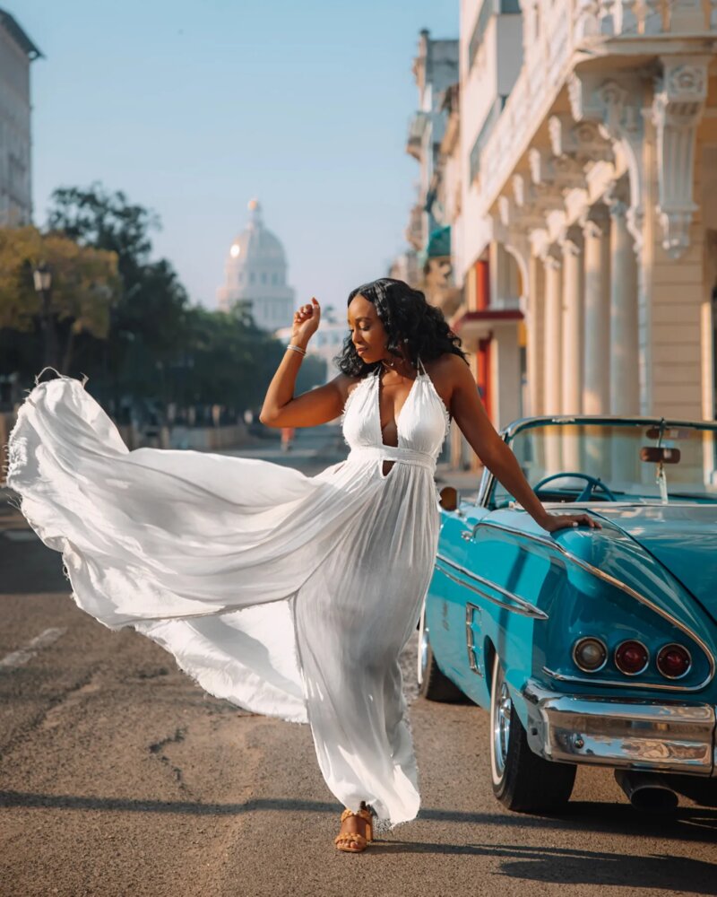 Zee James, co-founder of Voyagers Escape, wears white dress blowing in the breeze next to a vintage turquoise convertible in Cuba