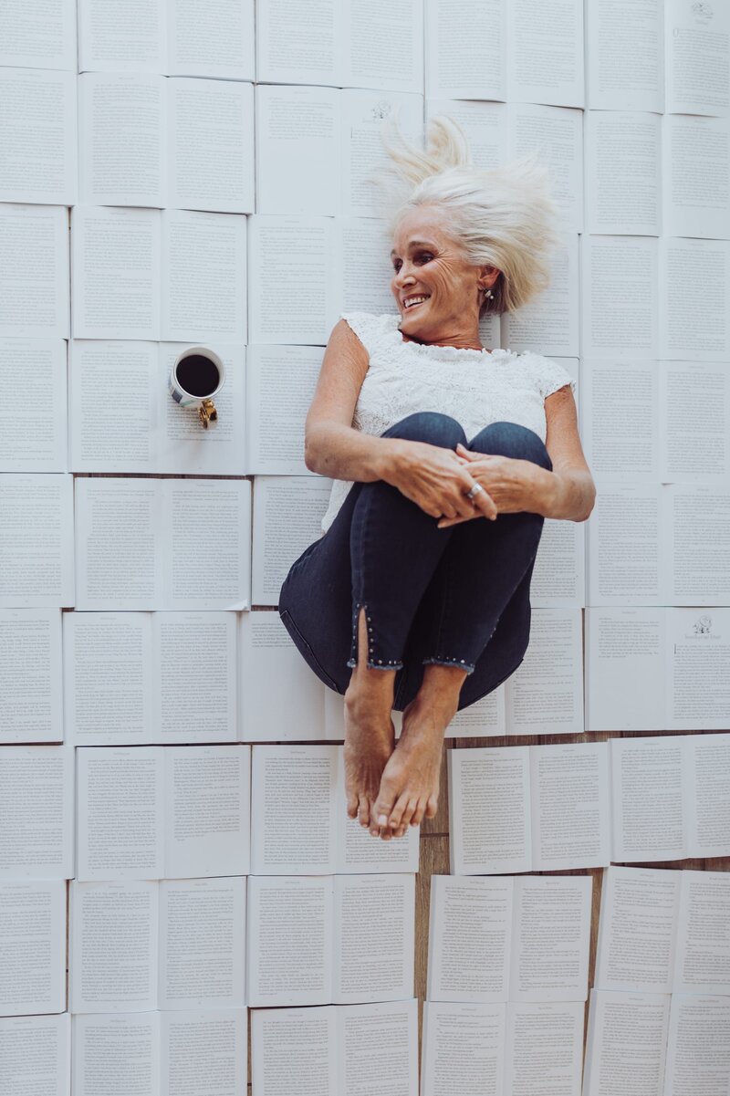 Woman wearing a white top, jeans lays barefoot while curled in a ball atop a bunch of open books. She looks over to a cup of black coffee laying beside her.